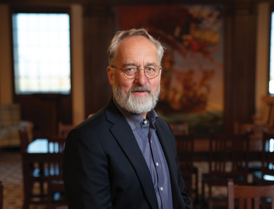 Man with gray beard and glasses wearing dark blazer over blue shirt stands in room with wooden furniture and windows