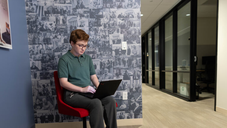 Person seated on red chair typing on laptop beside wall of black-and-white photo collage