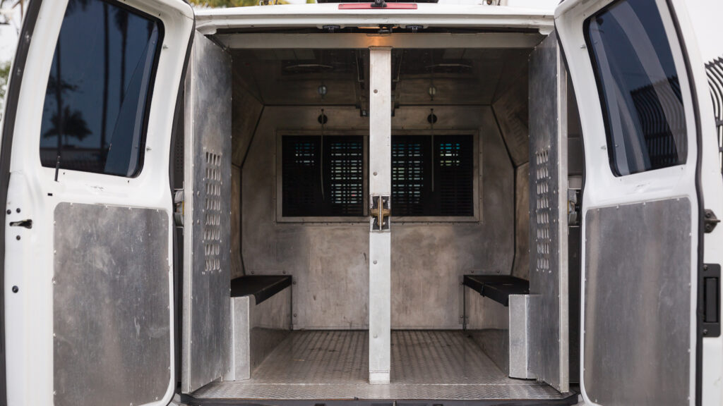 Rear doors open on a police van, showing metal bench seats used for transporting prisoners
