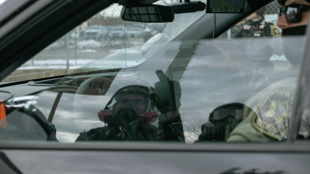 Person wearing a gas mask sits inside a vehicle near law enforcement officers at a protest scene