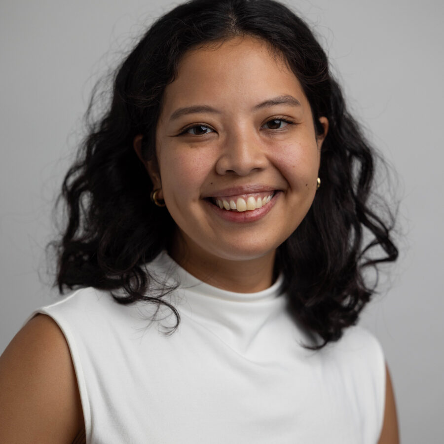 Woman with dark wavy hair and gold hoop earrings smiling in white shirt against gray background.