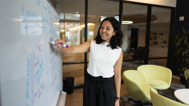 Woman with shoulder-length dark hair writing on whiteboard with purple marker in modern office with yellow chairs