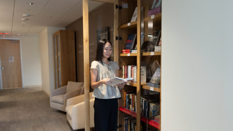 Woman reading an open book while standing beside a wooden bookcase in an office with beige armchairs