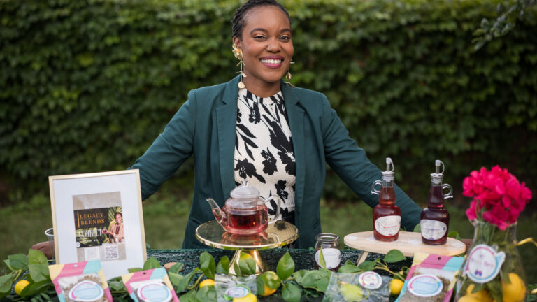 Woman smiling behind table of Legacy Blends teas, syrups, and jams outdoors