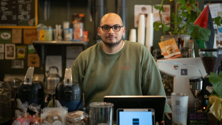 Man in green Habaka Cafe sweatshirt standing behind counter with coffee equipment and pastries