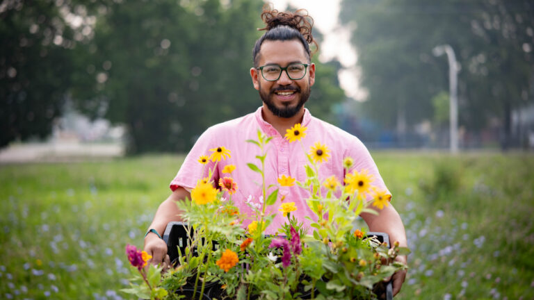 Smiling man in pink shirt holds crate of colorful flowers in a grassy field
