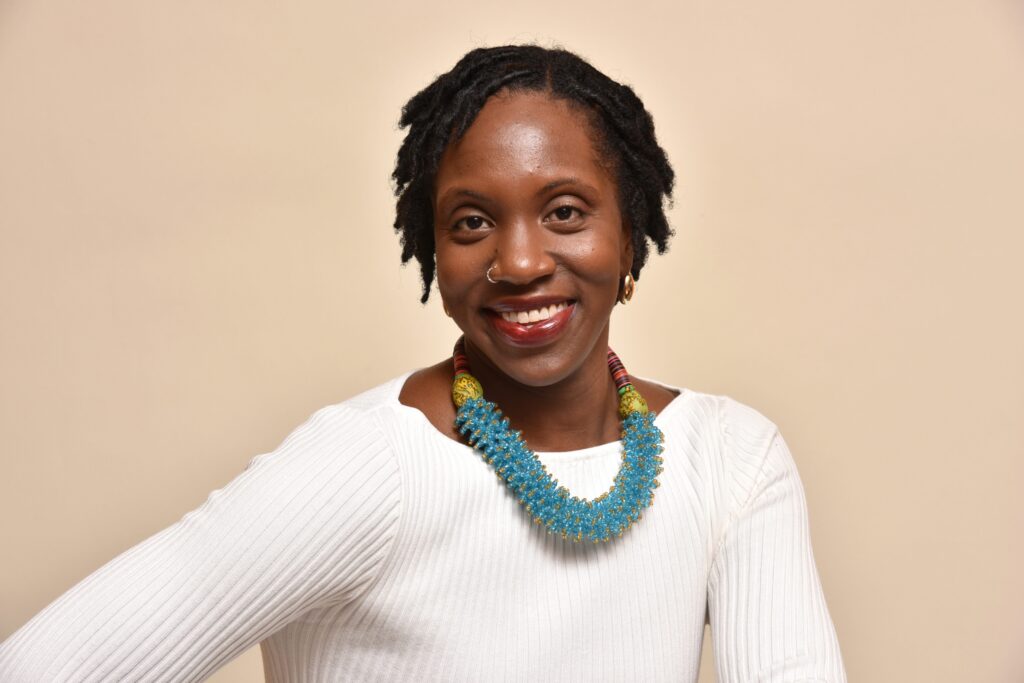 Woman with locs and a beaded blue necklace smiles in a studio headshot