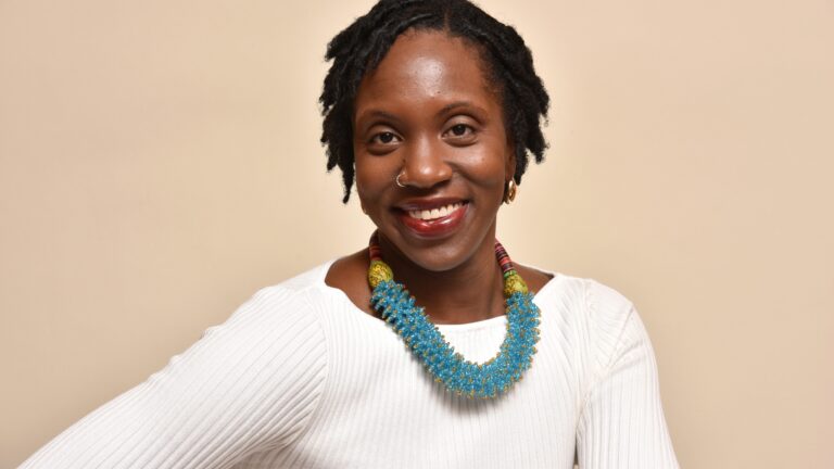 Woman with locs and a beaded blue necklace smiles in a studio headshot