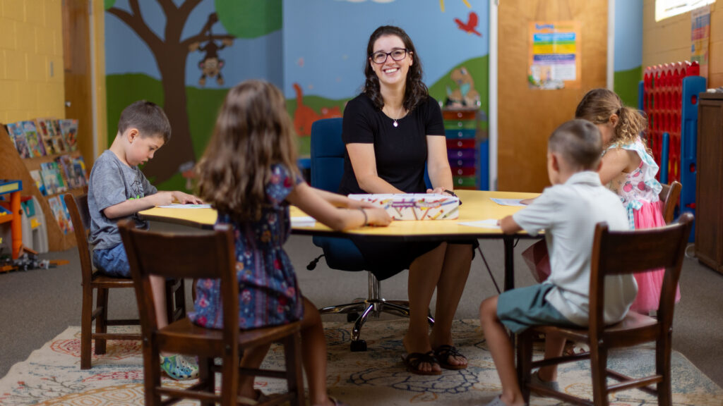 Katy Duckstein smiles at a table with children coloring in a classroom setting