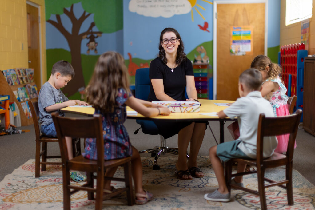Katy Duckstein smiles at a table with children coloring in a classroom setting