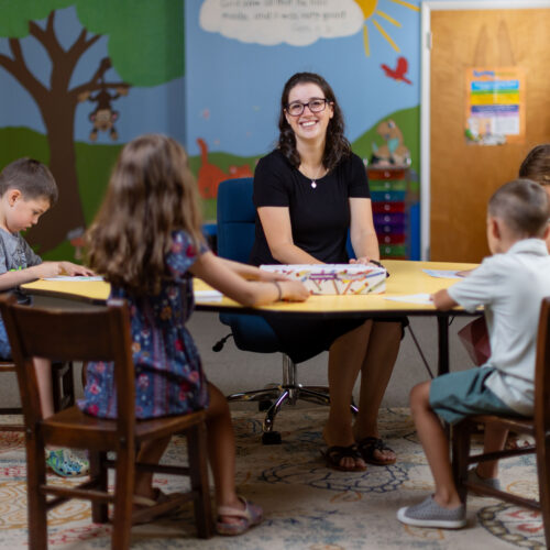 Katy Duckstein smiles at a table with children coloring in a classroom setting