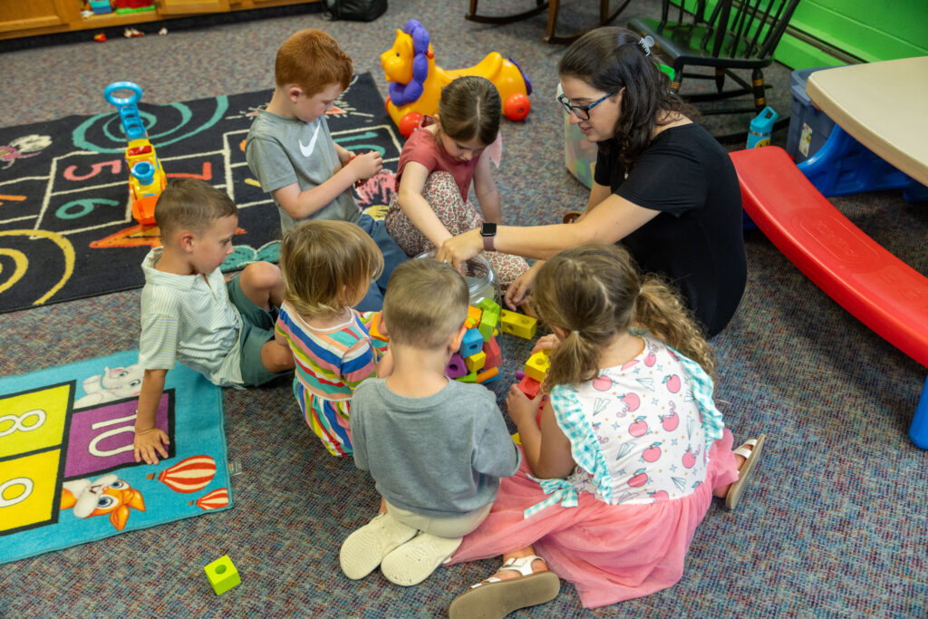Children sit on a classroom rug with a teacher, sorting colorful blocks together