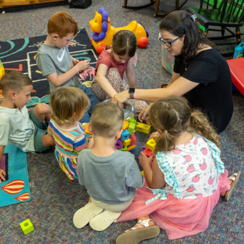 Children sit on a classroom rug with a teacher, sorting colorful blocks together