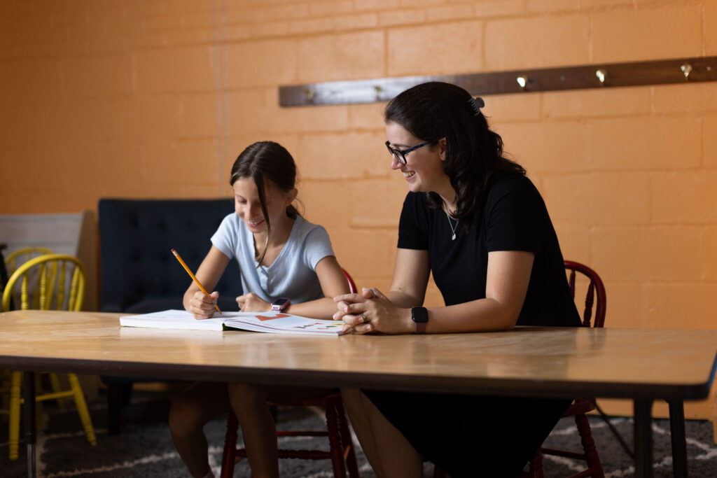 Woman smiles beside a girl writing in a workbook at a table