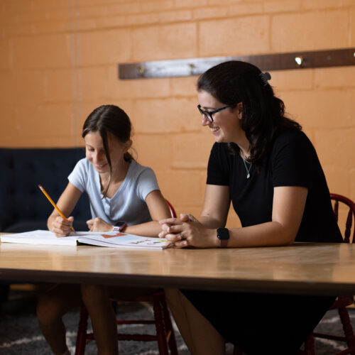 Woman smiles beside a girl writing in a workbook at a table