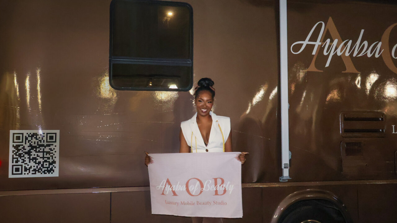 Brandi smiling, holding an Ayaba of Beauty sign in front of her mobile beauty salon