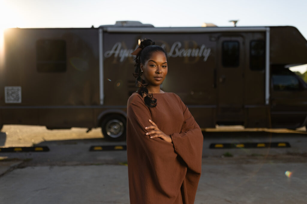 Woman with arms crossed stands before a mobile beauty salon truck labeled Ayana Beauty