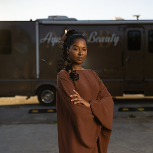Woman with arms crossed stands before a mobile beauty salon truck labeled Ayana Beauty