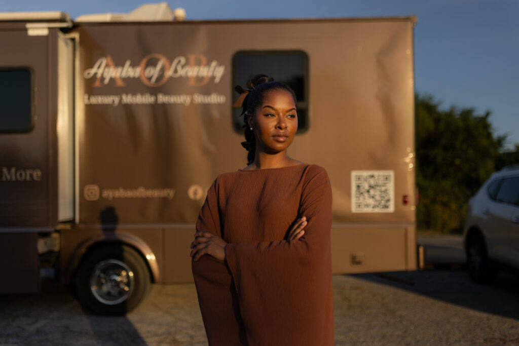 Woman with arms crossed stands in front of the Ayaba of Beauty mobile beauty studio truck