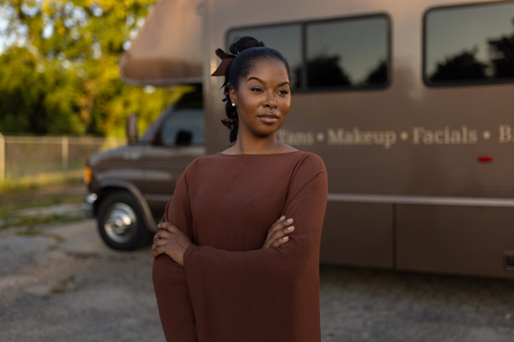 Woman stands arms crossed in front of a mobile beauty services van offering tans, makeup, and facials