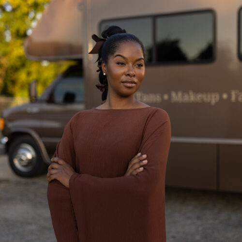 Woman stands arms crossed in front of a mobile beauty services van offering tans, makeup, and facials