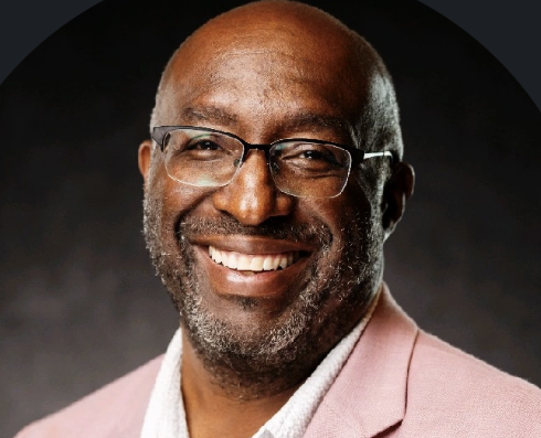 Bald man with glasses and beard smiling in pink blazer and white shirt
