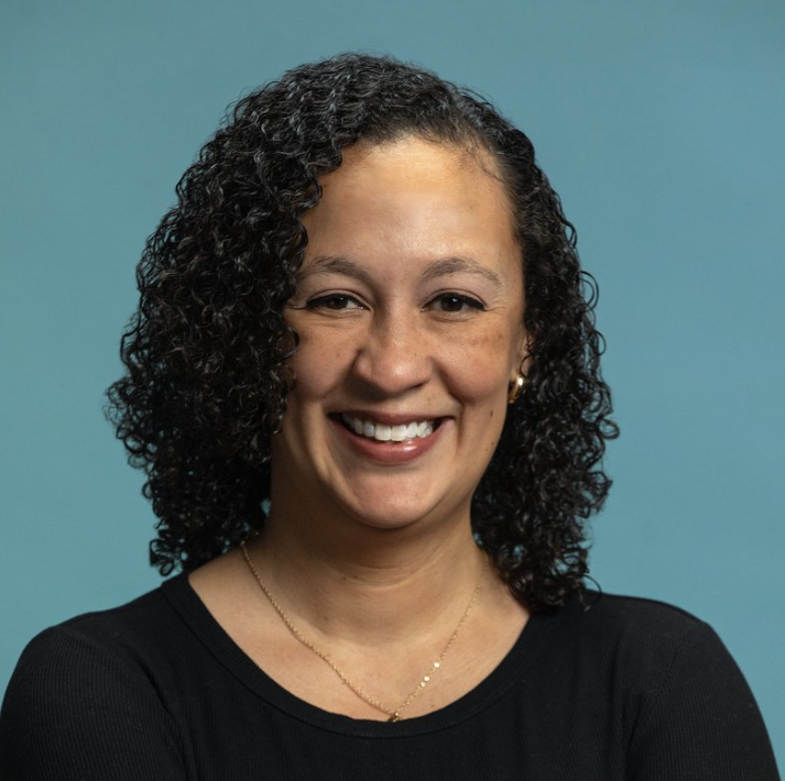 Woman with curly hair smiles in black top, arms crossed, against blue background