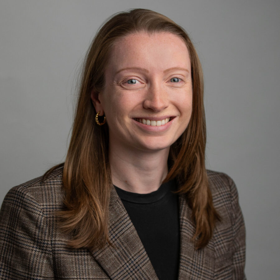 Woman with long hair smiling in a plaid blazer, professional headshot against gray background