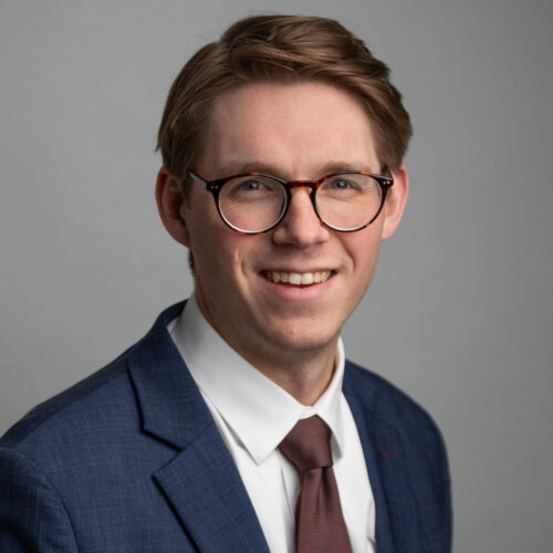 Headshot of a man in glasses and a navy suit smiling against a gray background