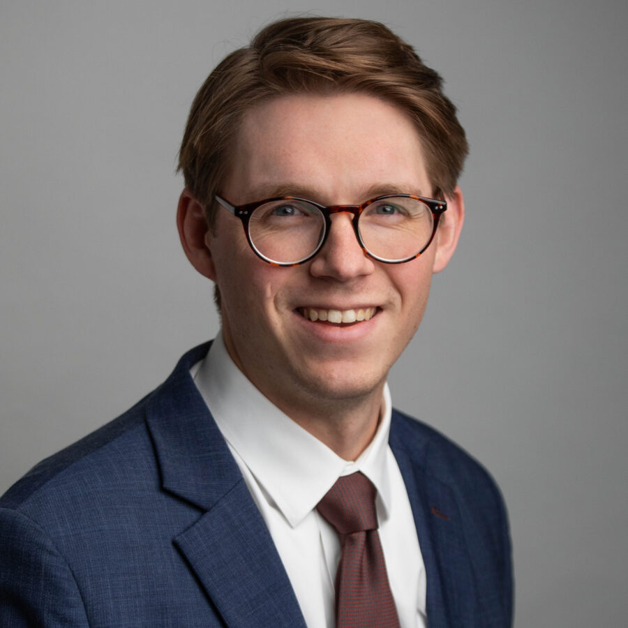 Headshot of a man in glasses and a navy suit smiling against a gray background
