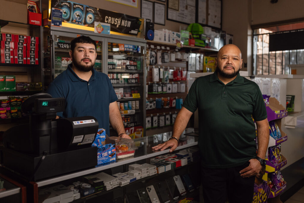 Two men stand behind convenience store counter near phone accessories and tobacco displays