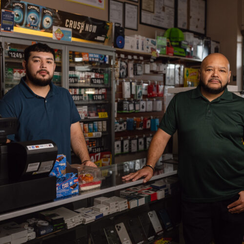 Two men stand behind convenience store counter near phone accessories and tobacco displays