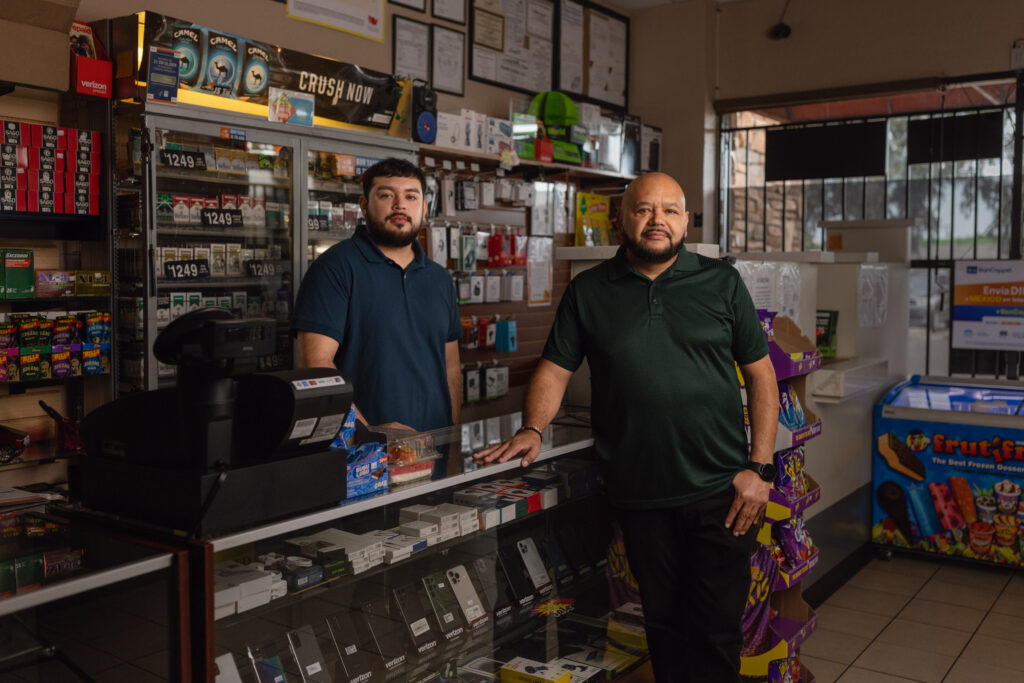 Two men stand behind the counter of a convenience store filled with phones and accessories