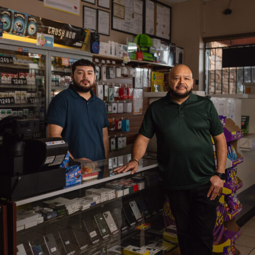 Two men stand behind the counter of a convenience store filled with phones and accessories