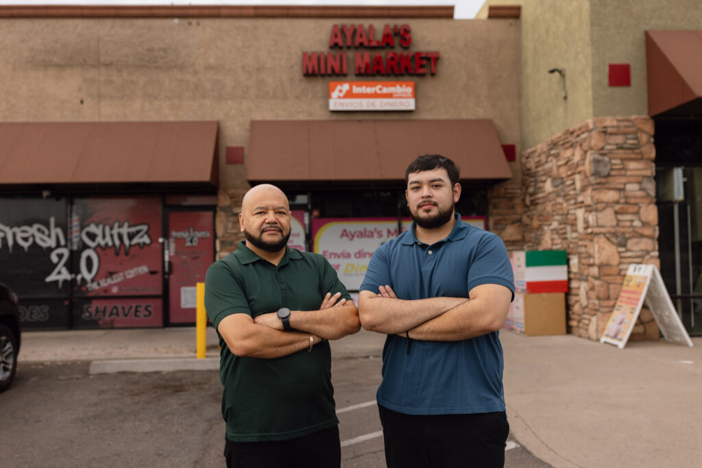 Two men stand arms crossed outside Ayala's Mini Market, a storefront with an InterCambio money transfer sign