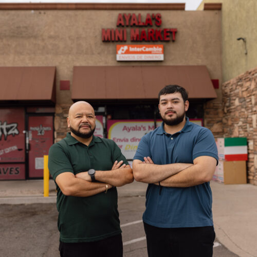 Two men stand arms crossed outside Ayala's Mini Market, a storefront with an InterCambio money transfer sign