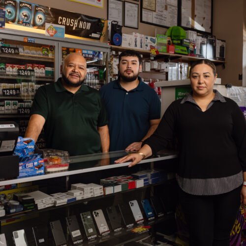 Three people stand behind a convenience store counter lined with phones and cigarettes