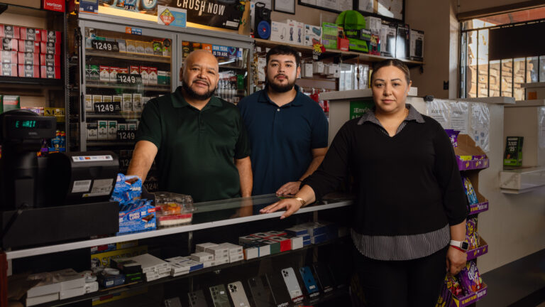 Three people stand behind a convenience store counter lined with phones and cigarettes