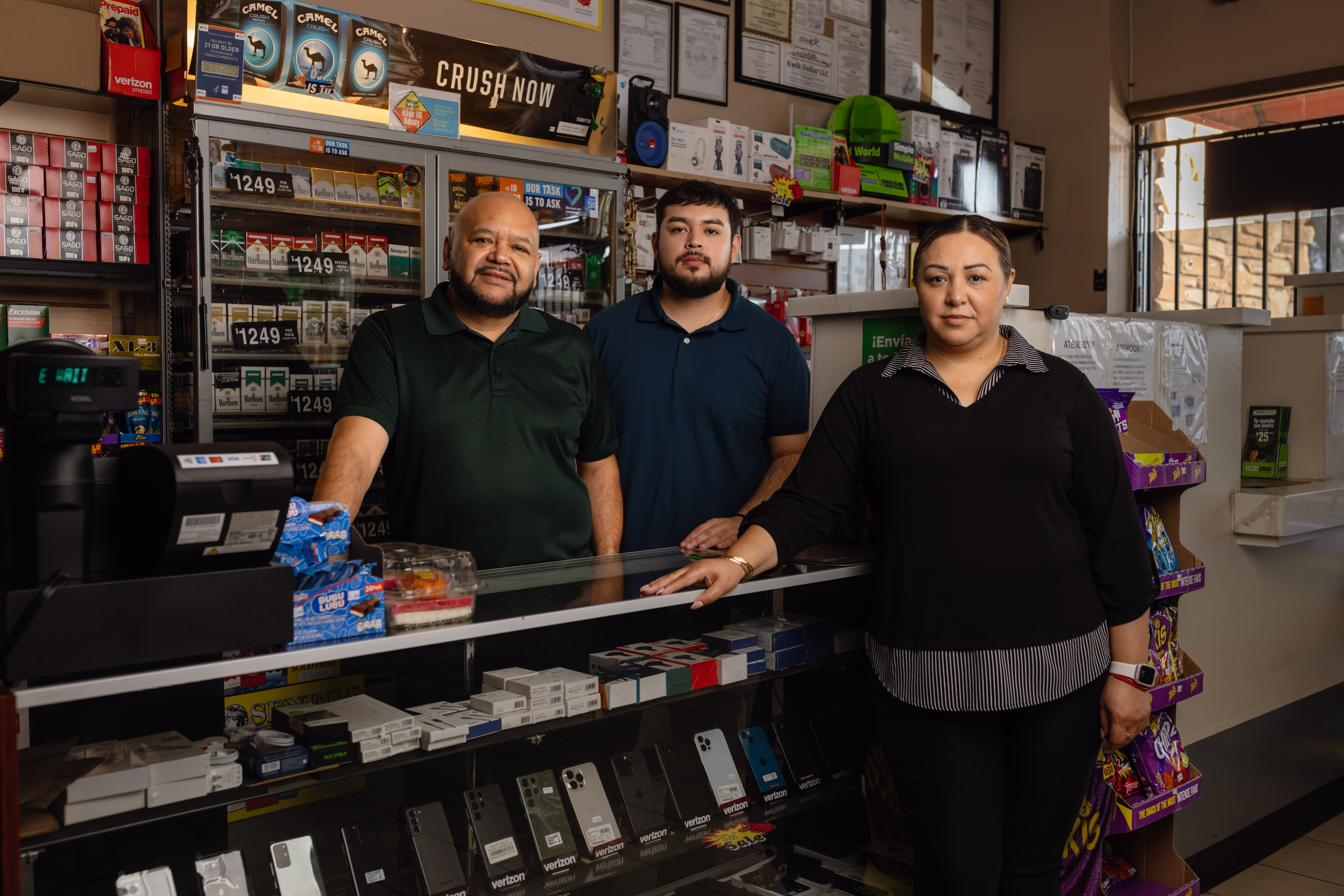 Three people stand behind a convenience store counter lined with phones and cigarettes