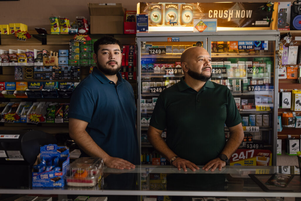 Two men stand behind a convenience store counter near a cigarette display case