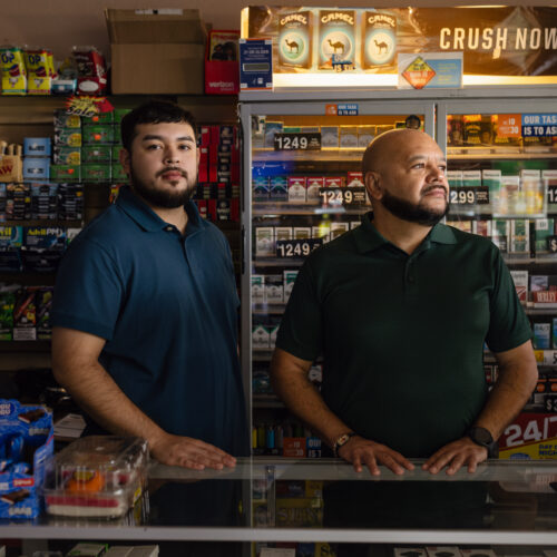 Two men stand behind a convenience store counter near a cigarette display case