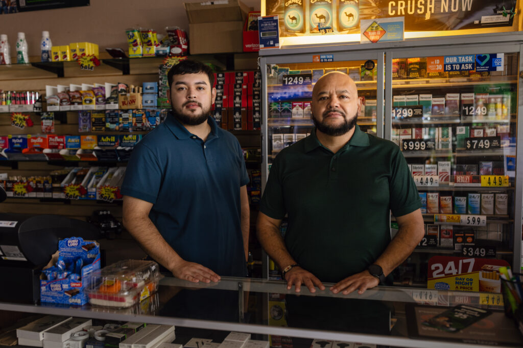 Two men stand behind a convenience store counter lined with tobacco products and price signs