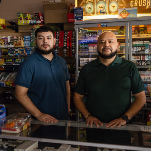 Two men stand behind a convenience store counter lined with tobacco products and price signs