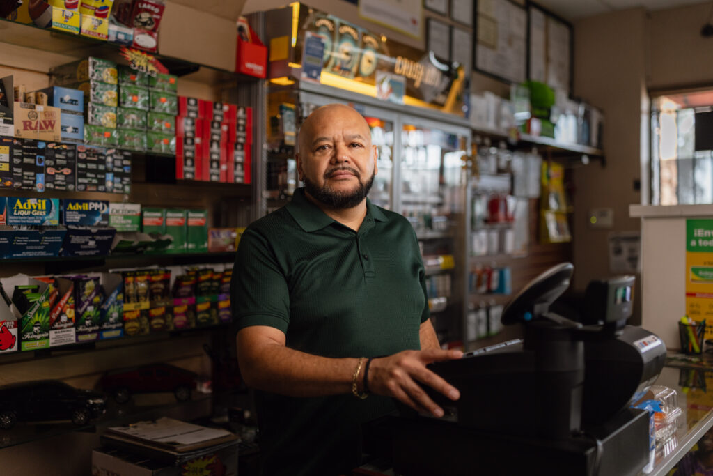 Man standing behind counter of convenience store shelves stocked with tobacco products and medicine