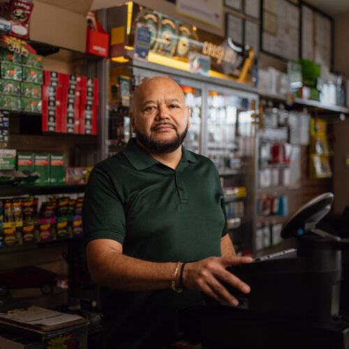 Man standing behind counter of convenience store shelves stocked with tobacco products and medicine
