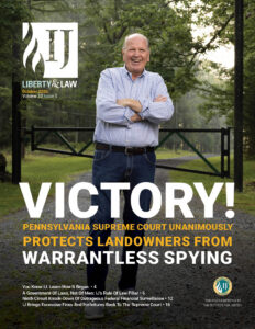 Man stands smiling with arms crossed on gravel driveway, gated wooded property behind him