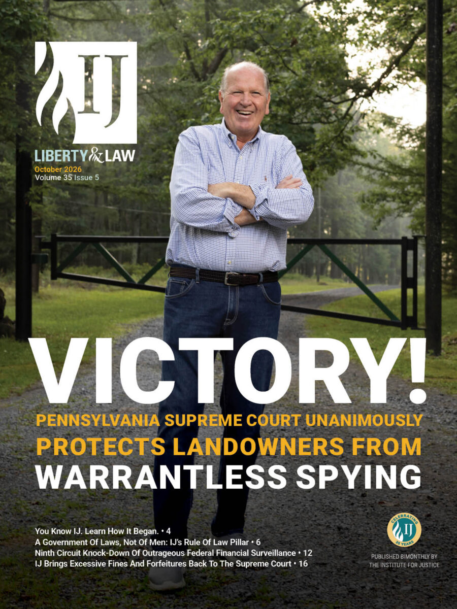 Man stands smiling with arms crossed on gravel driveway, gated wooded property behind him