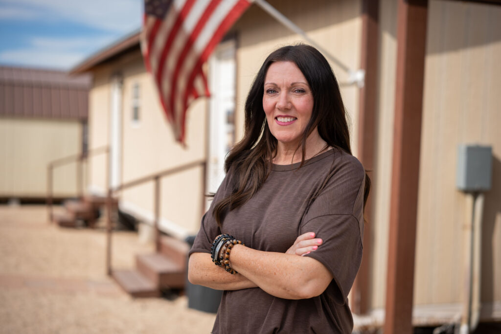 Woman with arms crossed smiles outside a manufactured home with an American flag