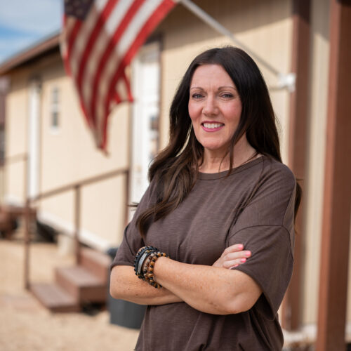 Woman with arms crossed smiles outside a manufactured home with an American flag