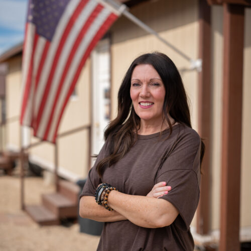 Woman with arms crossed smiling outdoors near an American flag in Pinal, Arizona